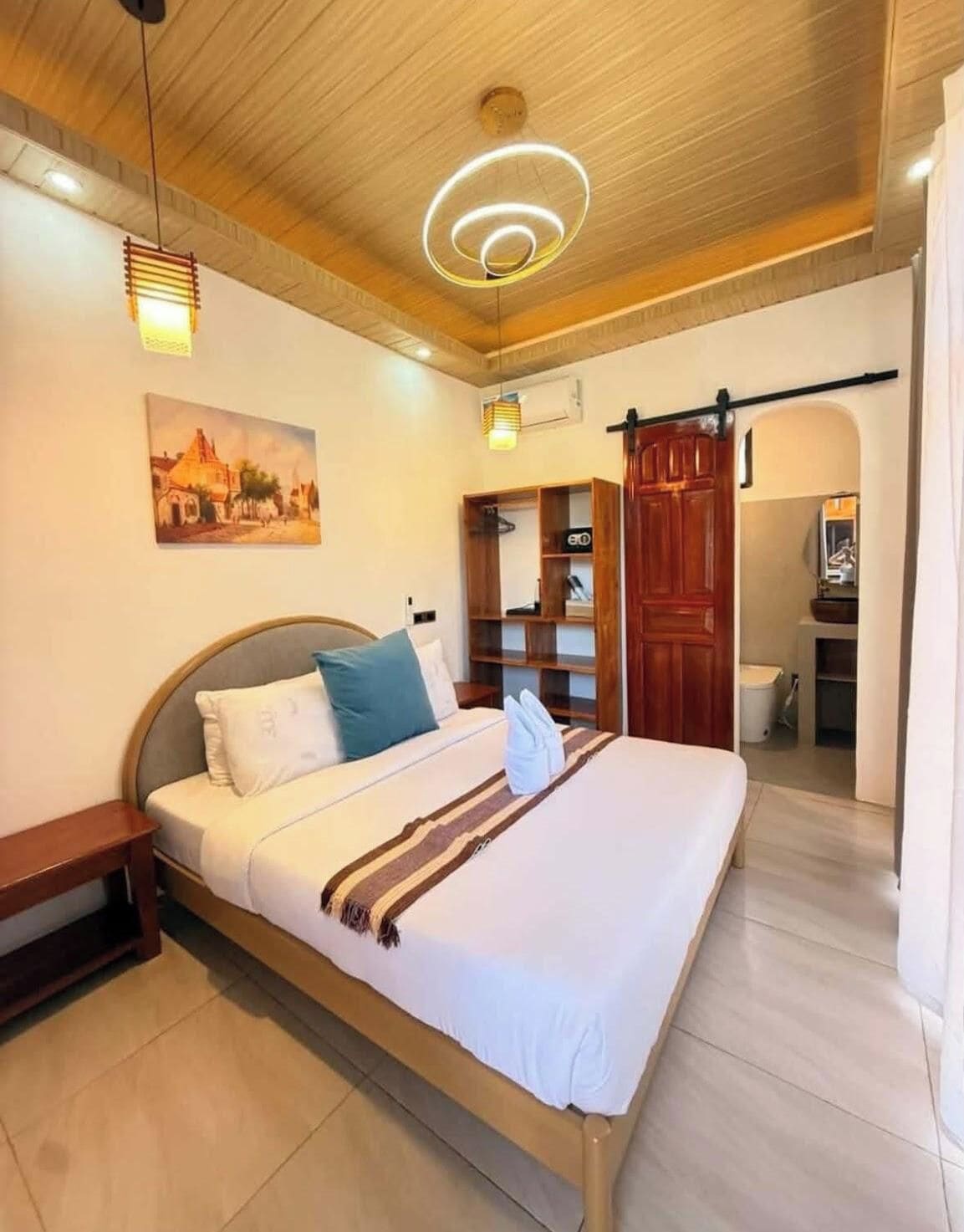 Une chambre d’hôtel avec un lit, un plafond en bois, un tableau accroché au mur, une armoire et une salle de bain visible à travers une arche.