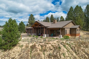 A two-story log cabin with a covered porch, surrounded by a desert landscape with sparse vegetation and tall pine trees. - The Cascade Ranch House | 24 Acres of Idaho Mountain Country (Cascade)