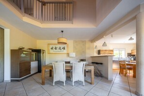 Un espace repas doté d’un haut plafond et d’un escalier. On y trouve une table à manger en bois avec des chaises, un luminaire suspendu et un tableau encadré au mur.