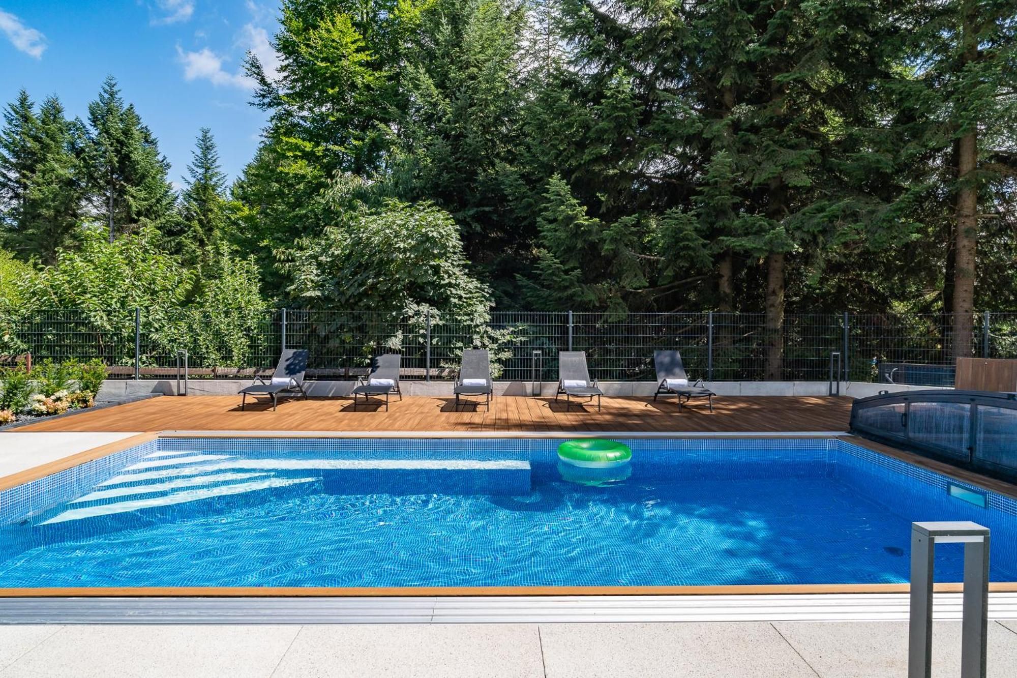A swimming pool with lounge chairs and a wooden deck, surrounded by a metal fence and trees.