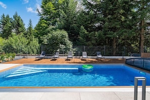 A swimming pool with lounge chairs and a wooden deck, surrounded by a metal fence and trees.