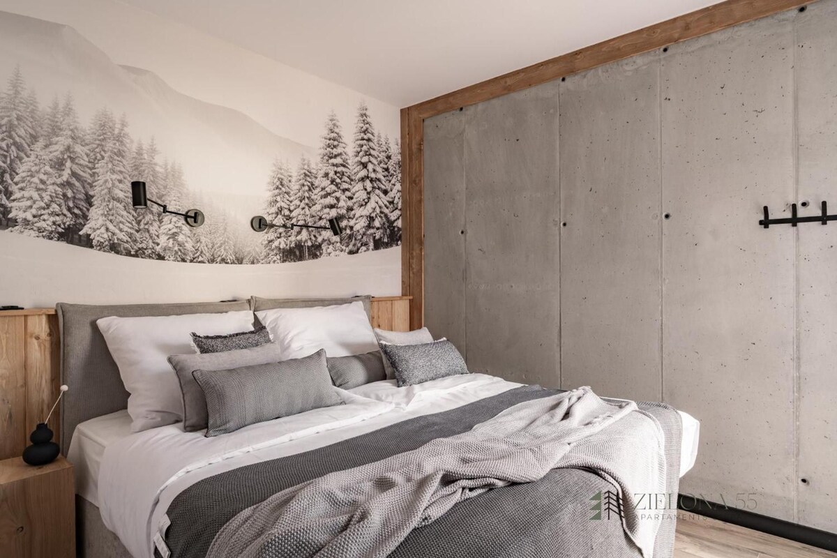 A bedroom with a bed, pillows, a blanket, a nightstand, a lamp, a wardrobe, and a wall with a forest mural.
