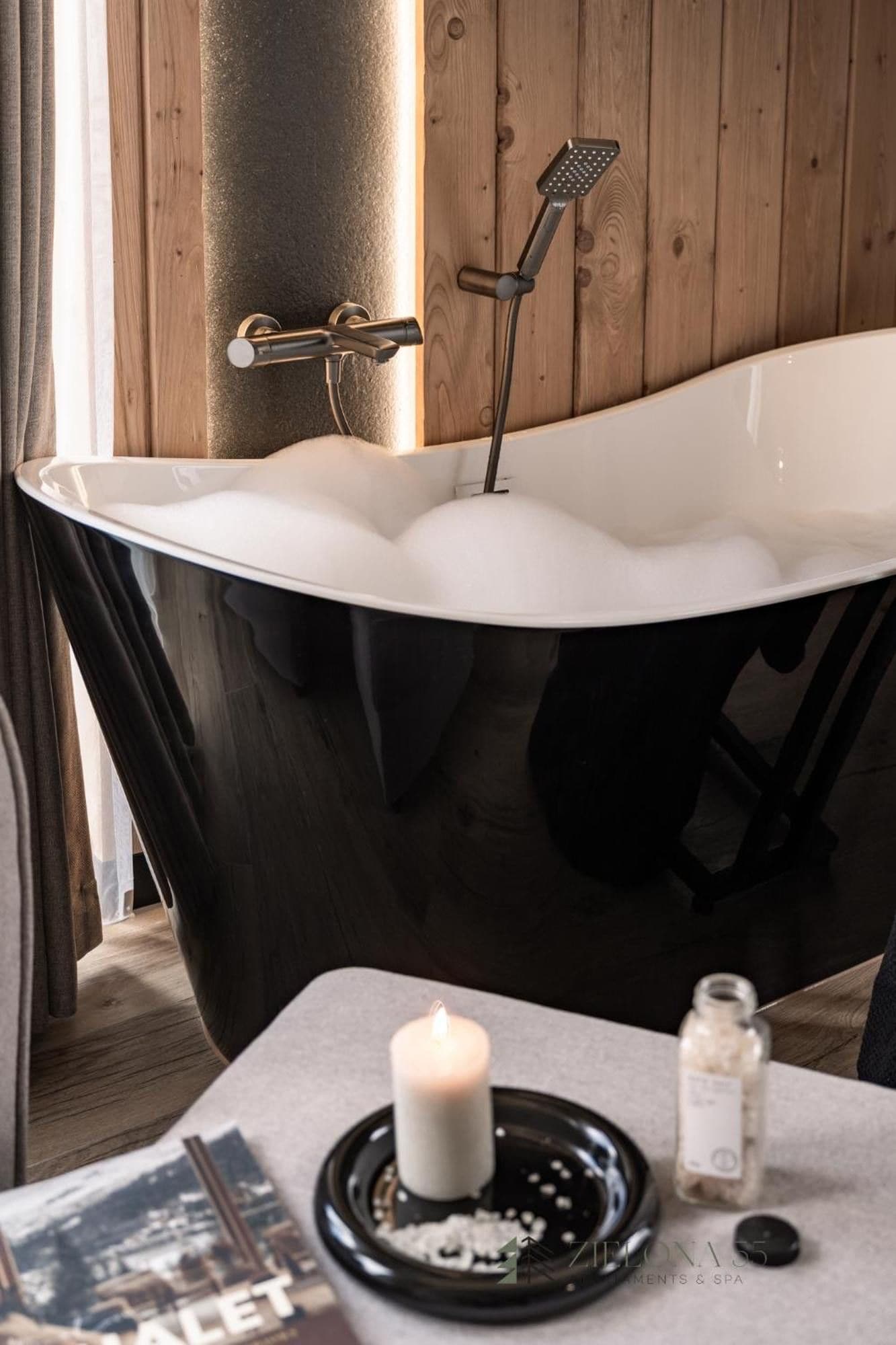 Een moderne badkamer met een vrijstaand bad, een kaars en een kleine fles op een tafeltje.
