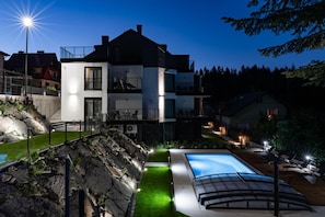 Un edificio moderno de dos pisos con piscina, iluminación exterior y jardín ajardinado.