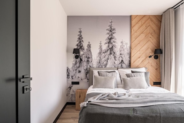 Un dormitorio con cama, cabecera de madera, mesita de noche, lámpara de pared y ventana con cortinas.
