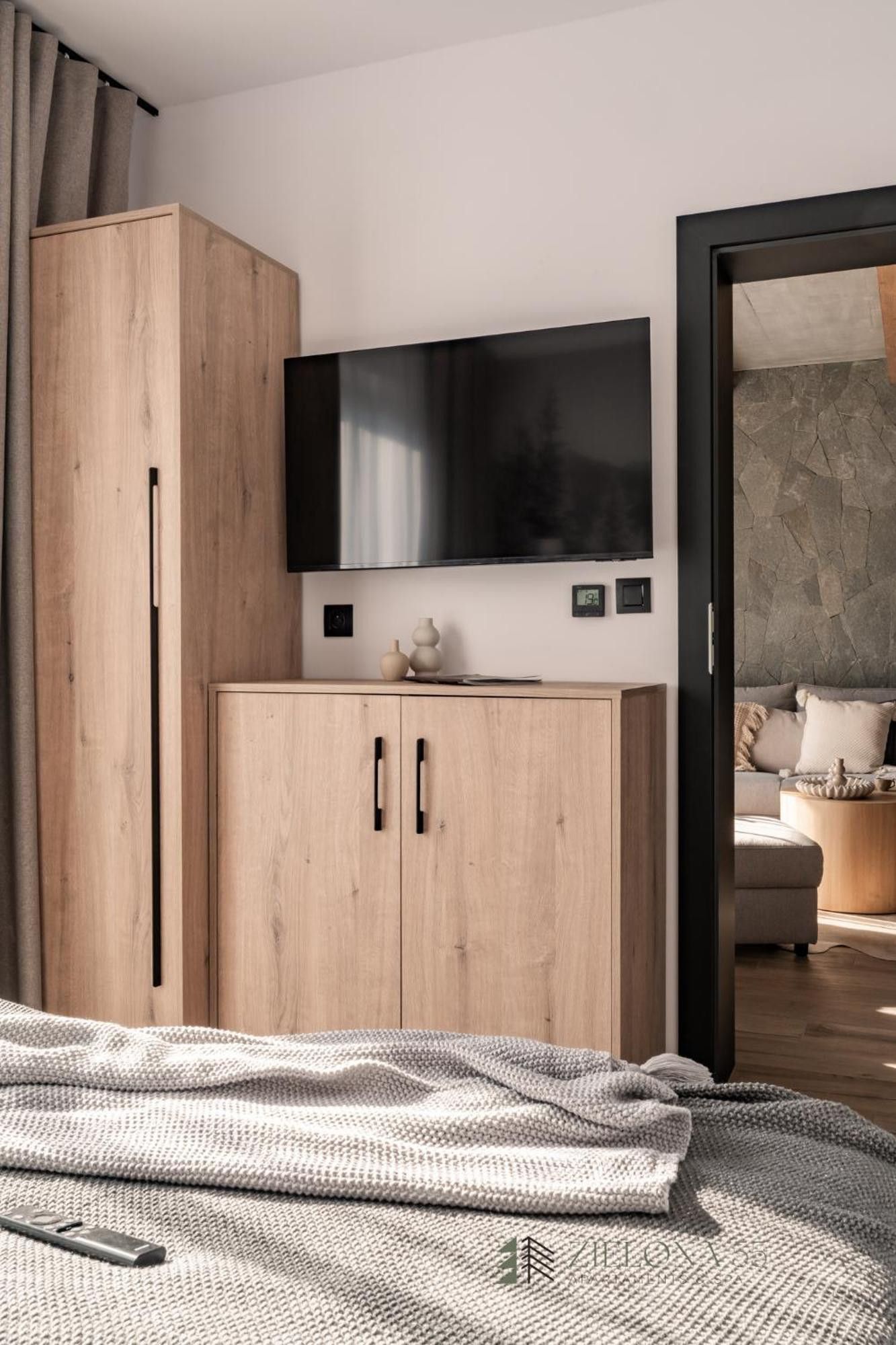 A modern bedroom with a wooden wardrobe, a flat-screen TV mounted above it, and a cozy bed with a knitted blanket.