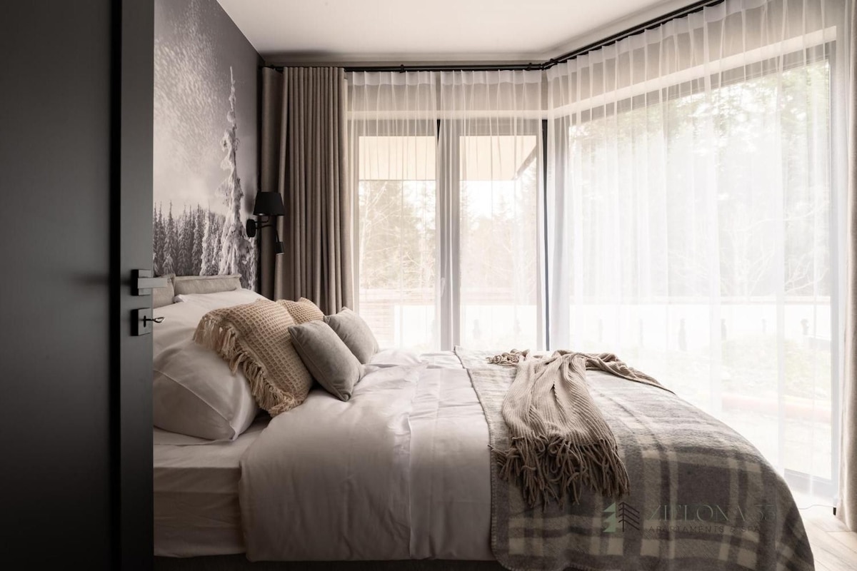 Une chambre avec un grand lit, une vue sur les arbres à travers de fines rideaux, et une lampe fixée au mur.