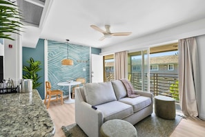 A modern living room with a grey sofa, a dining table with chairs, a ceiling fan, and a view of a residential area through large windows. - The Blue Lagoon - in Pacific Beach w/ comm pool (San Diego)