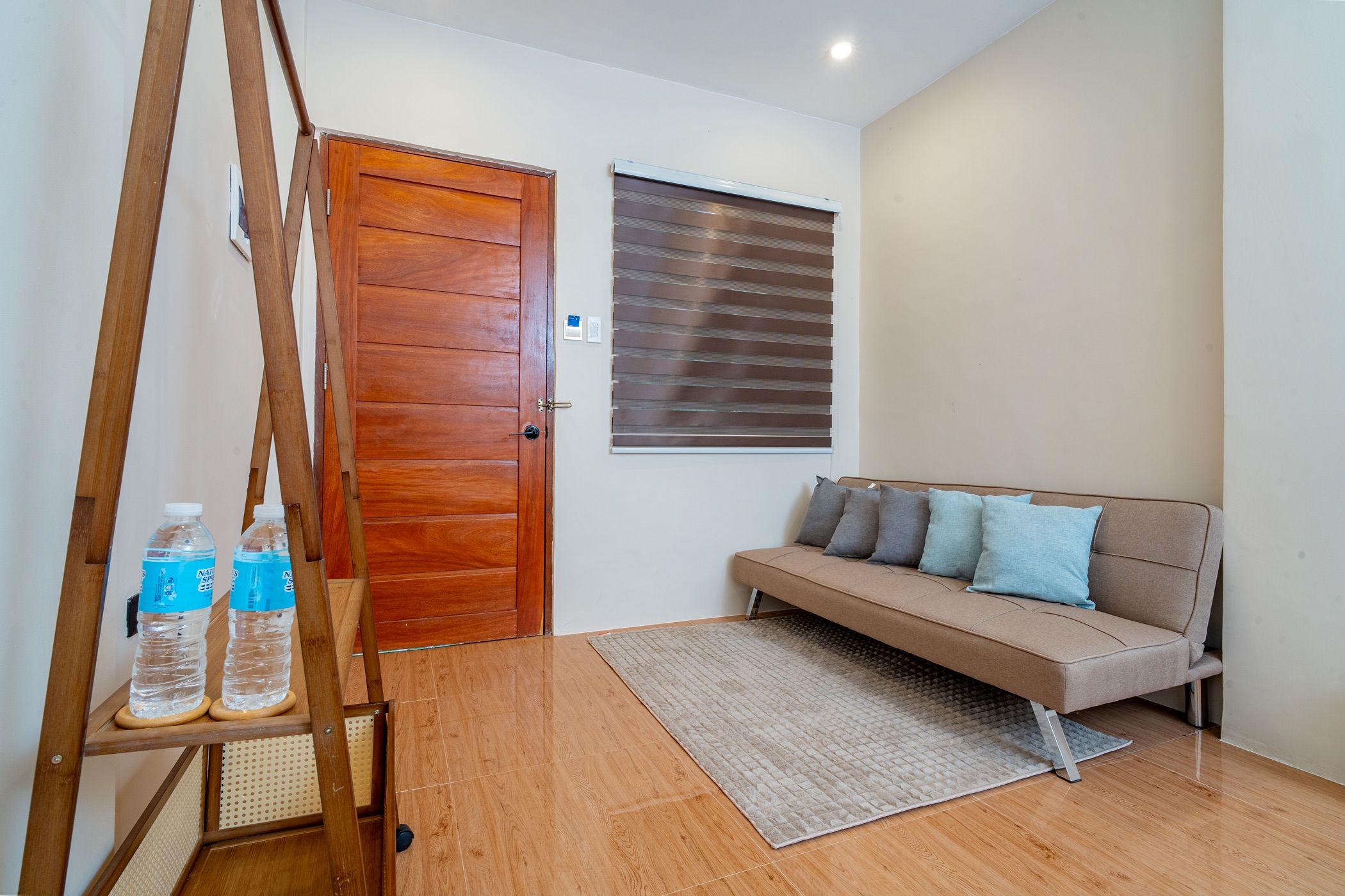 Um quarto com porta de madeira, um sofá com almofadas, piso de madeira e uma janela com persianas.