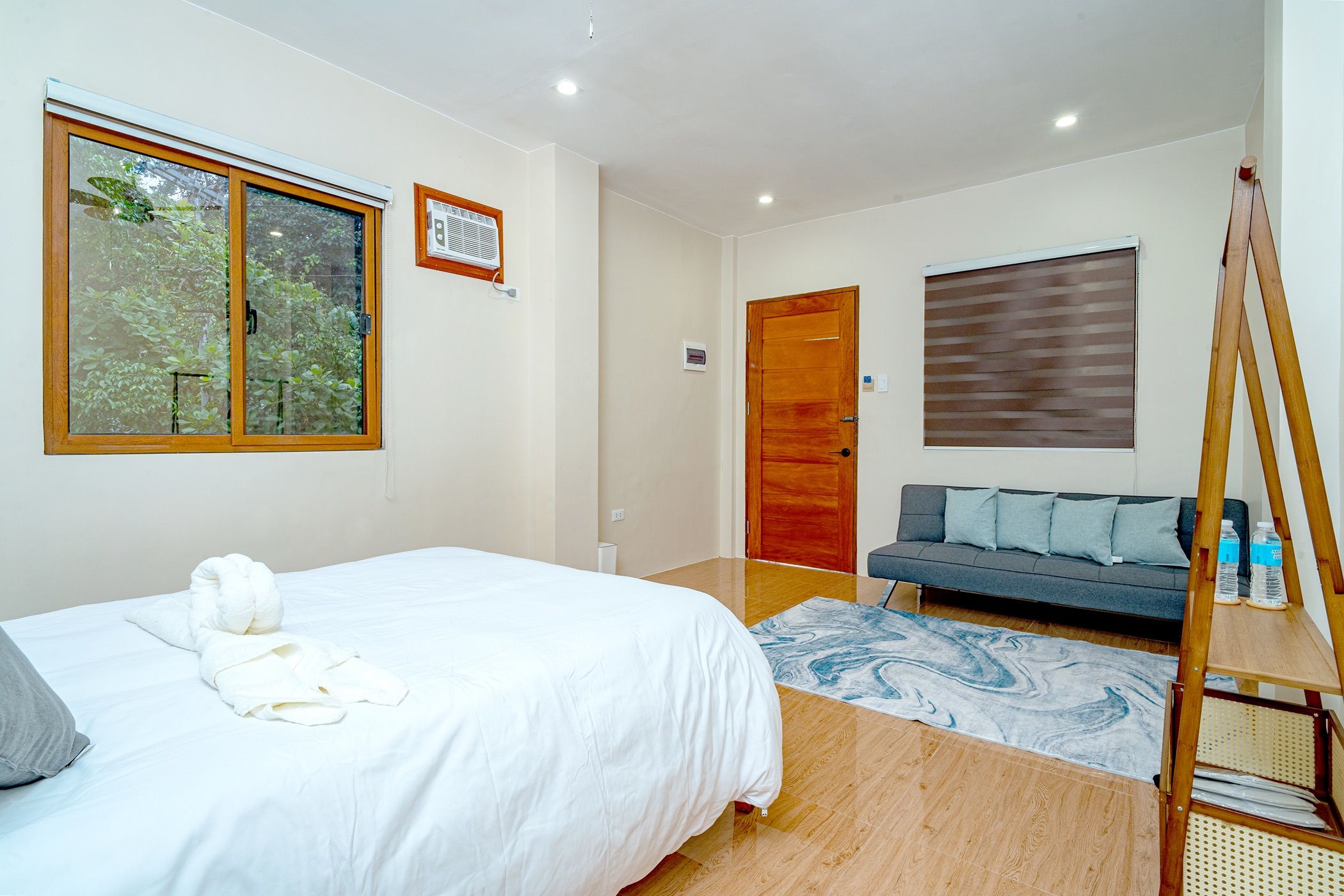 Quarto com cama, sofá, piso de madeira e janela com vista para árvores.