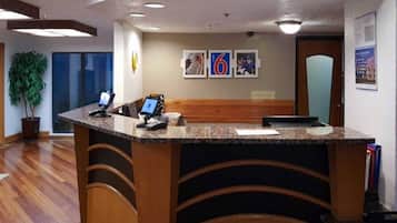 A reception desk with a computer, a phone, and a seating area. There are framed pictures on the wall and a plant to the left.