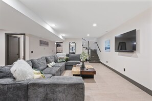 A modern living room with a grey sectional sofa, a coffee table, and a flat-screen TV mounted on the wall. - New! Sapphire Collection Lake View Lodge, Hot Tub & Fire Pit! (Somers)