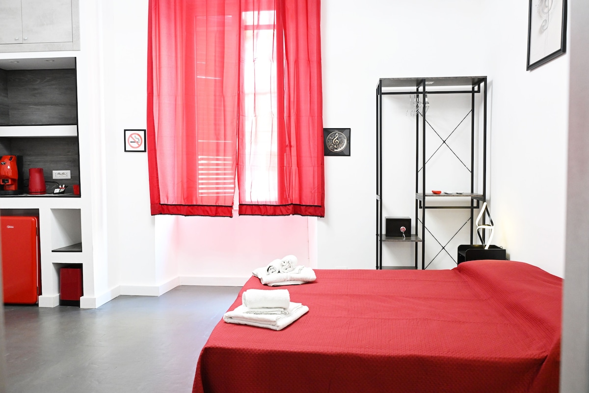 A hotel room with a bed, a red curtain, a shelf with toiletries, and a small red appliance.