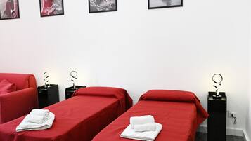 Una camera d'albergo con due letti, ciascuno con una coperta rossa e asciugamani bianchi. Sono presenti comodini neri con una lampada da comodino dal design moderno.