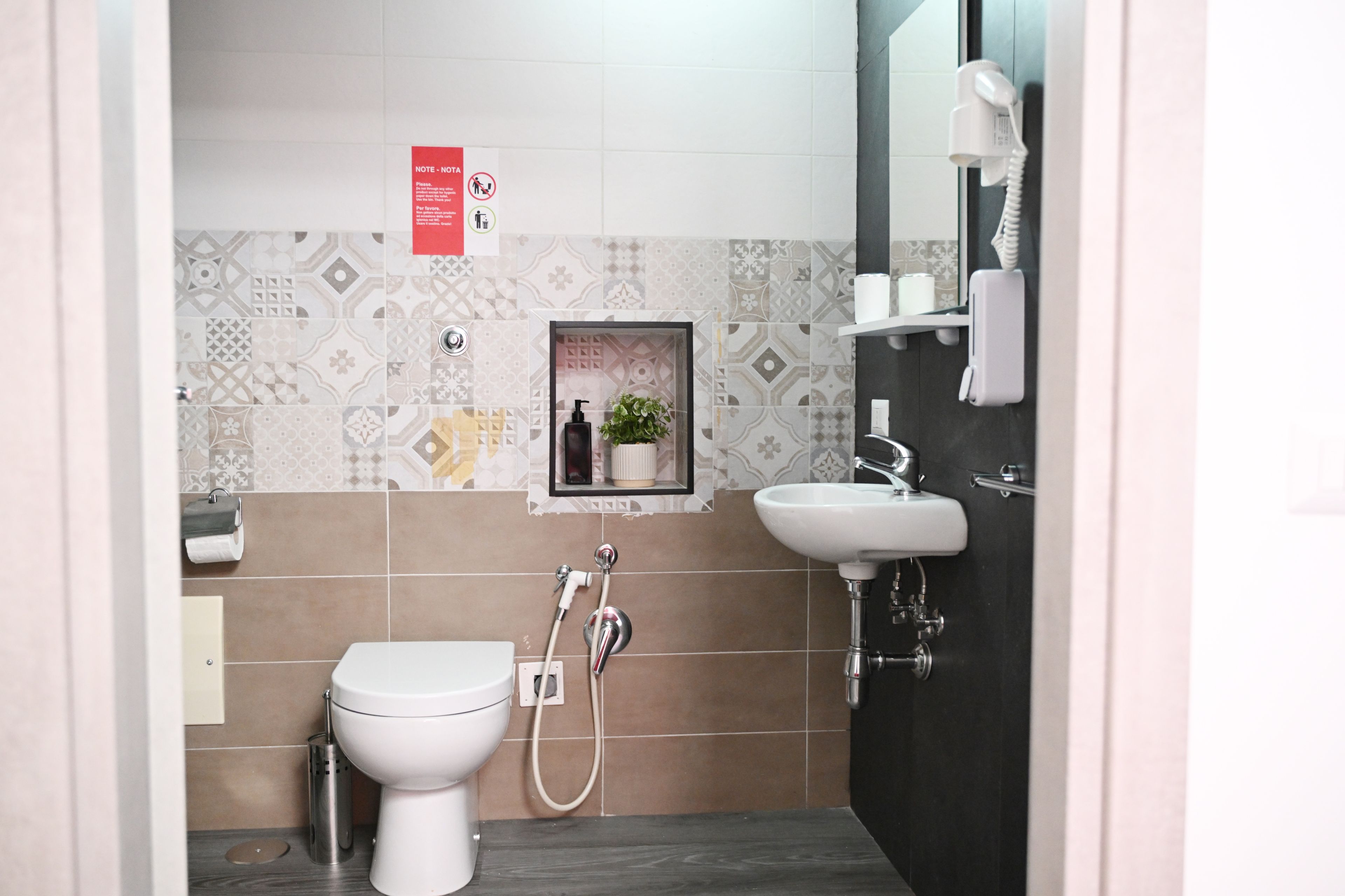Un bagno con toilette, bidet, lavandino e uno specchio. Si nota un cartello appeso al muro e un piccolo ripiano con una pianta.