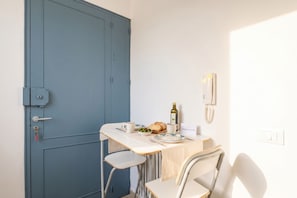Une pièce avec une porte bleue, une petite table recouverte d’une nappe blanche, une chaise et une bouteille d’huile d’olive.