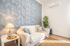 Quarto com papel de parede estampado, sofá branco, mesinha com abajur e uma planta.
