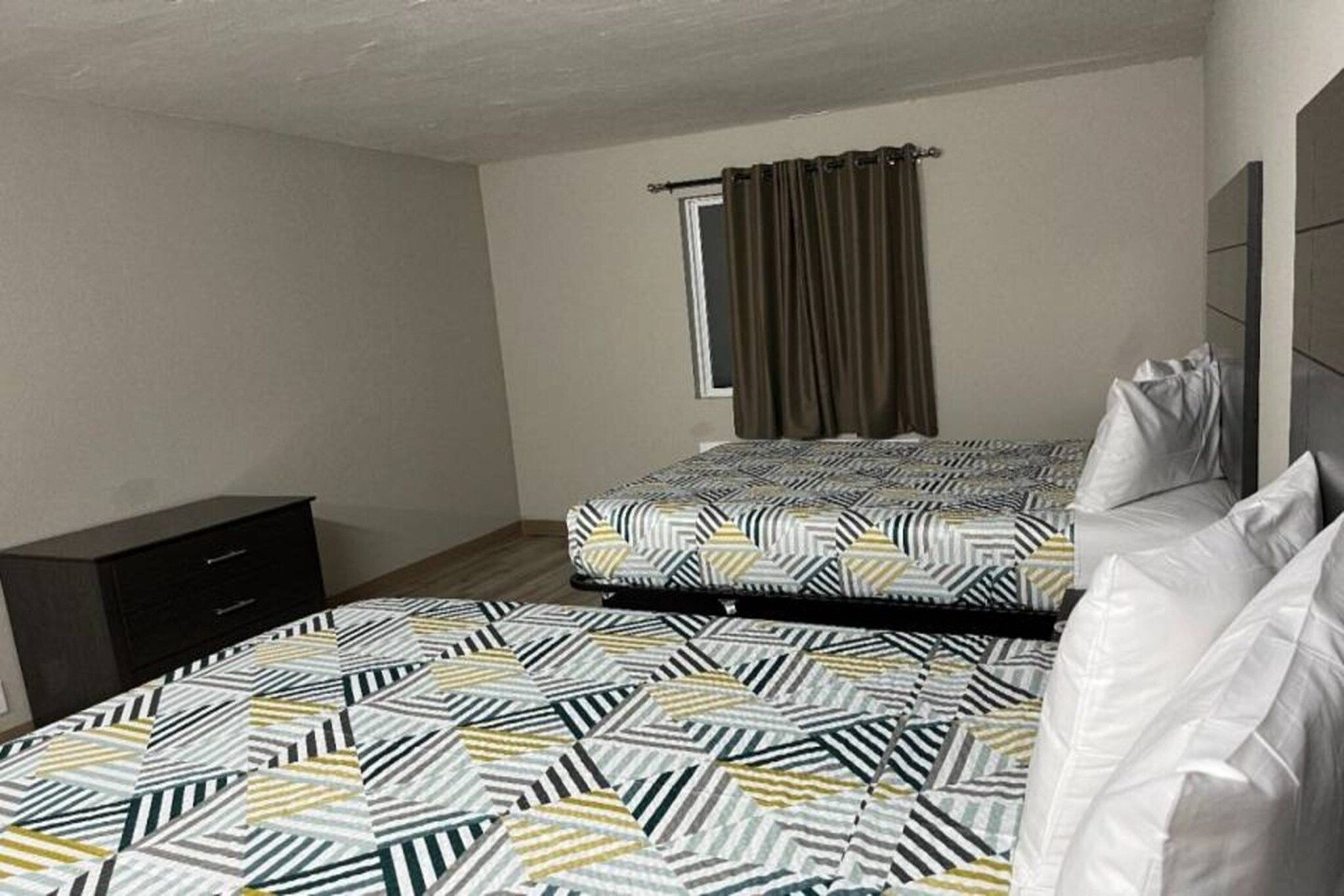A bedroom with two beds, a window with curtains, a dark dresser, and a wall-mounted headboard.