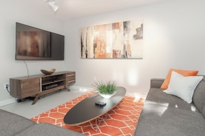 A modern living room with a flat-screen TV, a wooden entertainment unit, a grey sofa with an orange pillow, and a coffee table with a plant. - Riverside Home | Walk Downtown | Hot Tub | Firepit (Winter Park)