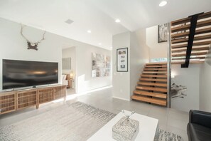 A modern living room with a flat-screen TV, wooden staircase, and artwork on the walls.