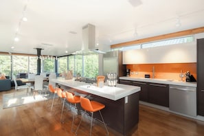A modern kitchen with a central island, bar stools, and a dining area with a view of the outdoors. - Riverside Home | Walk Downtown | Hot Tub | Firepit (Winter Park)