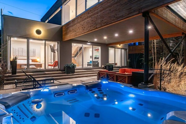 A modern outdoor hot tub area with a wooden deck, a staircase leading to a glass door entrance, and a seating area with red chairs.