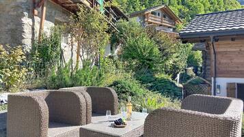 Gartenmöbel aus Rattan mit Tisch, Weinflasche und Gläsern, vor der malerischen Kulisse von Bergen und traditionellen Holzhäusern.
