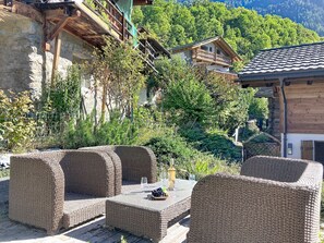 Gartenmöbel aus Rattan mit Tisch, Weinflasche und Gläsern, vor der malerischen Kulisse von Bergen und traditionellen Holzhäusern.