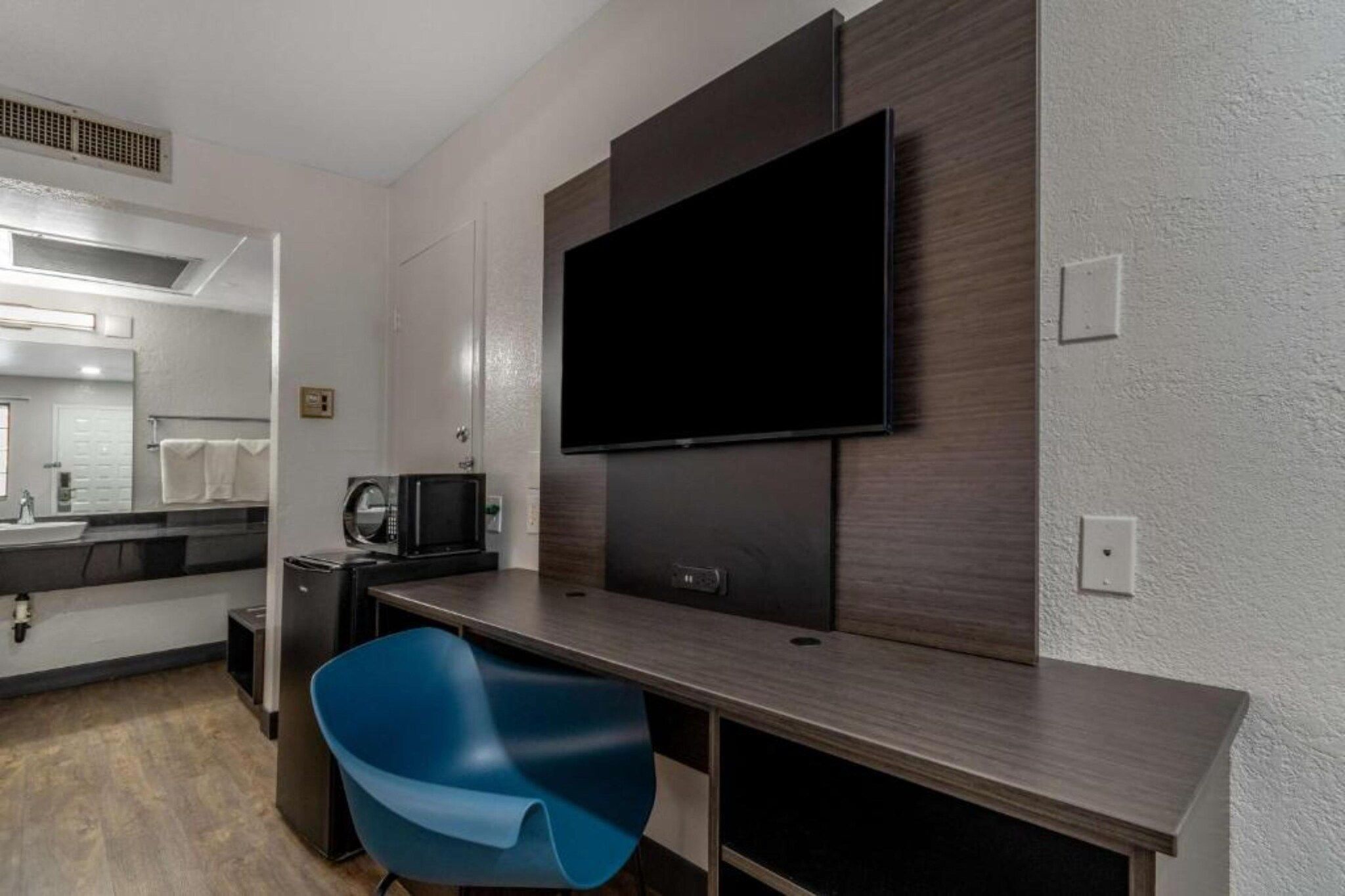 A modern hotel room with a flat-screen TV mounted on the wall, a small kitchenette, a blue chair, and a large mirror.