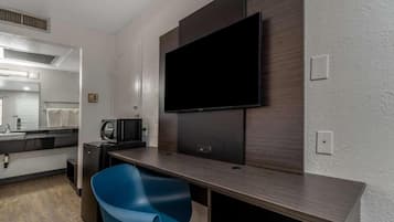 A modern hotel room with a flat-screen TV mounted on the wall, a small kitchenette, a blue chair, and a large mirror.