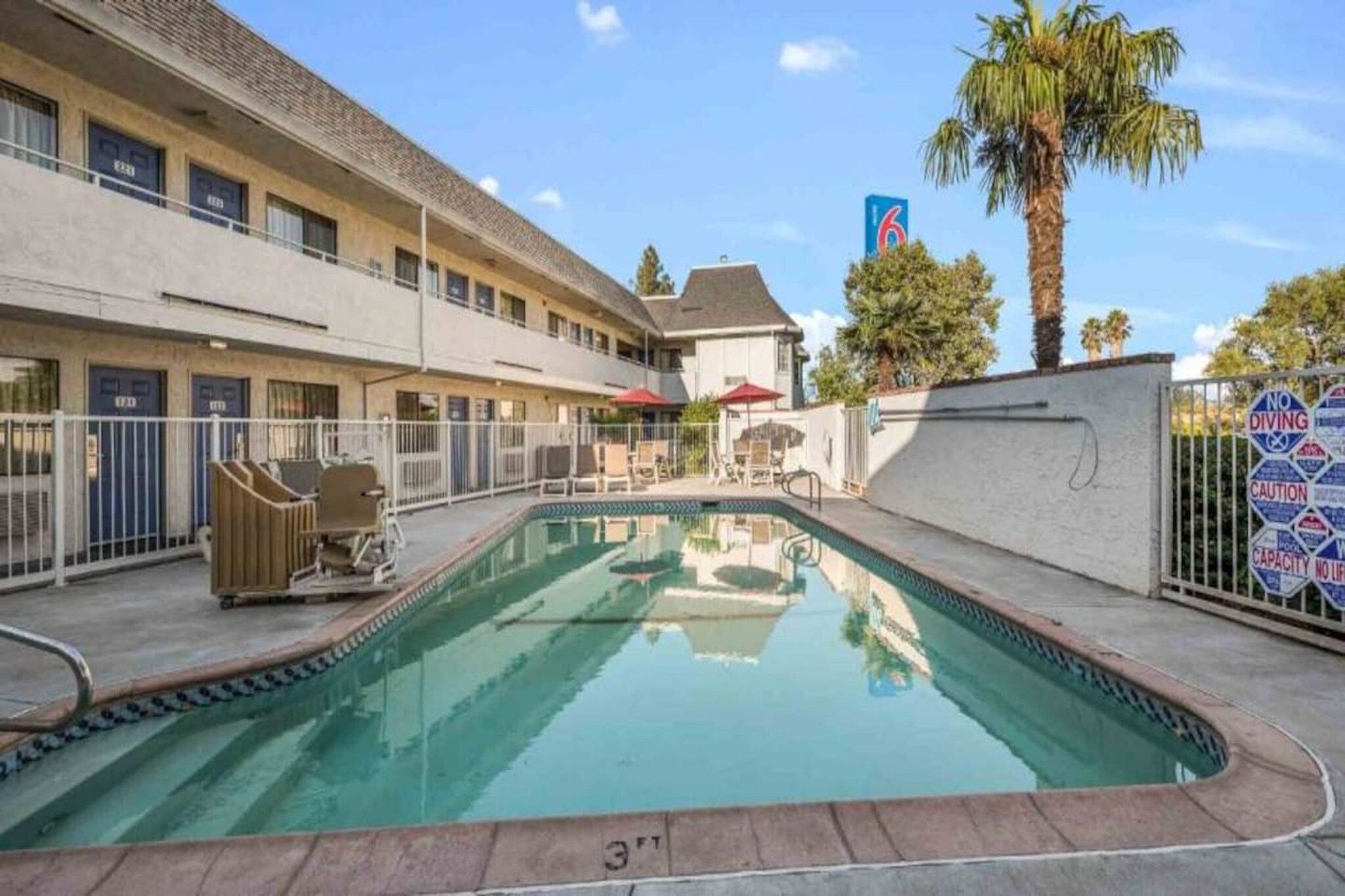 Une piscine entourée de chaises longues et de parasols, le tout bordé par un bâtiment doté de nombreuses portes et d’un palmier.