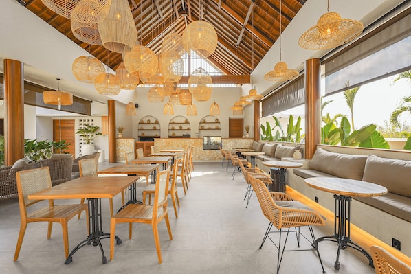 A spacious indoor area with wooden tables and chairs, a stone wall, and a large window overlooking greenery. - Siroco Hotel Uluwatu (Pecatu)