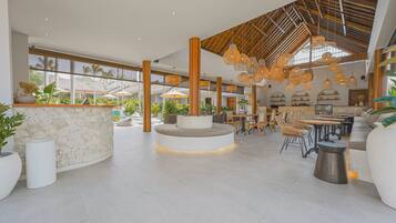 Un vaste espace intérieur doté d’un coin salon central, de plusieurs tables et chaises, le tout agrémenté de grandes fenêtres et de plantes.