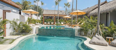 A swimming pool with lounge chairs and umbrellas, surrounded by palm trees and a thatched-roof building.