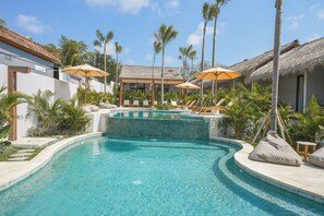 A swimming pool with lounge chairs and umbrellas, surrounded by palm trees and a thatched-roof building. - Siroco Hotel Uluwatu (Pecatu)