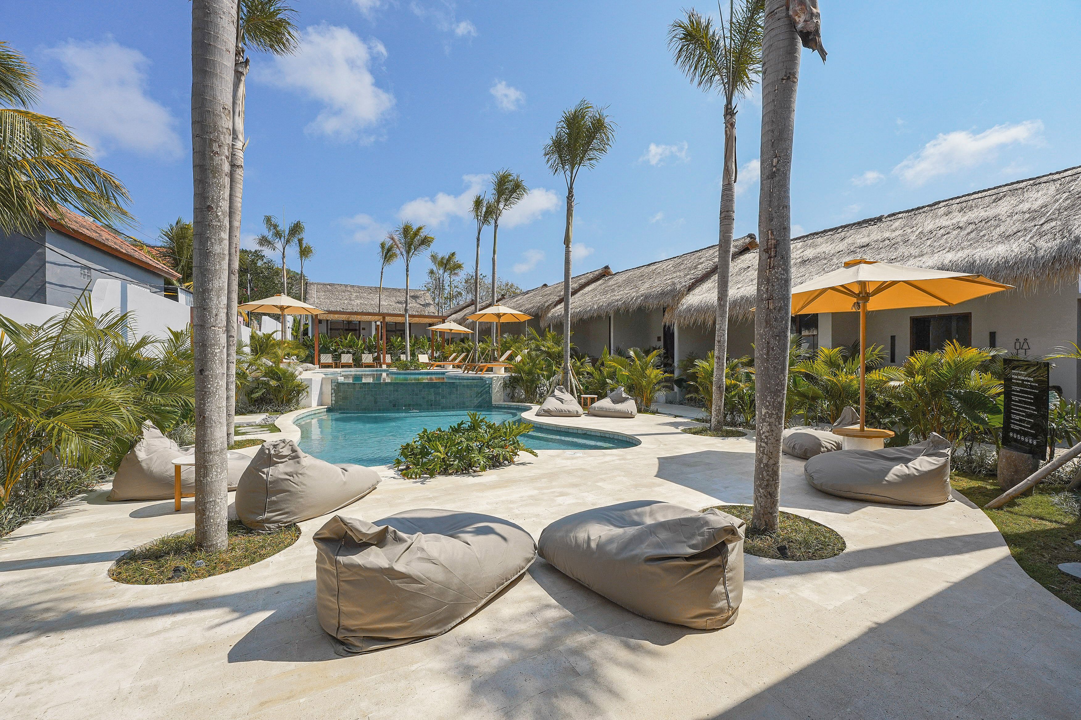 A pool area with lounge chairs and umbrellas, surrounded by palm trees and a thatched-roof building.