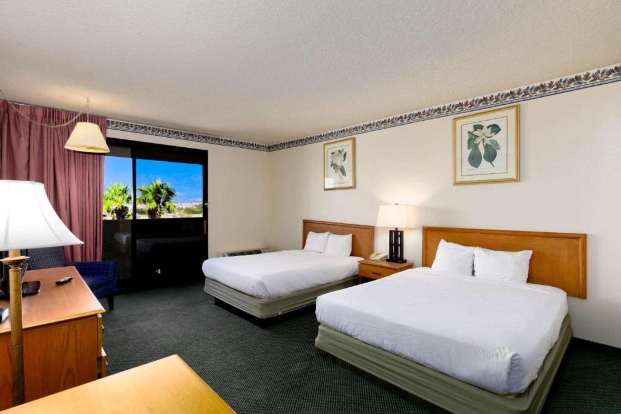 A hotel room with two beds, a desk, a chair, a lamp, and a view of palm trees through the window.