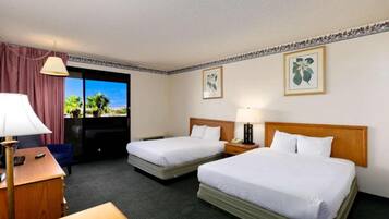 A hotel room with two beds, a desk, a chair, a lamp, and a view of palm trees through the window.