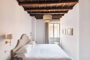 Habitació d'hotel amb sostre de fusta, llit amb roba de llit blanca, lampadeta de nit, llum penjada a la paret i finestra amb cortines.