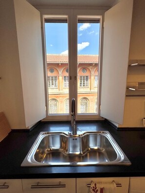 Cuina amb un rentaplatets d'acer inoxidable, una finestra que ofereix vista a un edifici amb finestres d'arc, i un taulell negre.