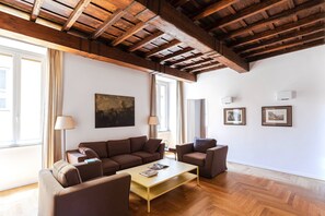 Un saló amb sostre de fusta, un sofà, una taula de centre i dos butaques. Hi ha dues finestres amb cortines, un quadre a la paret i dos imatges emmarcades.