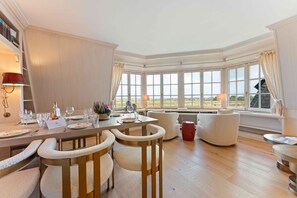 Un coin repas avec une table dressée pour quatre personnes, entourée de chaises rembourrées, d’un tabouret rouge et offrant une vue sur l’extérieur grâce à de grandes fenêtres.