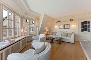 A room with a white sofa, two armchairs, a small round table, and a radiator. There are books on the radiator, a lamp on the table, and a window with curtains. - Twee Seen 33 - "Twee Seen 33" Vacation Rental (Kampen (Sylt))