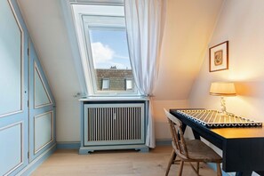 A room with a sloped ceiling, a radiator, a table with a patterned table runner, a chair, and a lamp. - Twee Seen 33 - "Twee Seen 33" Vacation Rental (Kampen (Sylt))