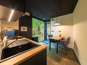 A modern kitchen with a sink, a dining area with a table and chairs, and a view of a forest through a glass door. - The Serene Escape - Olivenise Cabin (La Roche-en-Ardenne)