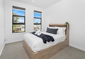 Un dormitorio con cama de madera, ropa de cama blanca, almohada a rayas negras y blancas y dos ventanas con vistas al exterior.