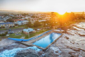 A swimming pool adjacent to a rocky shoreline with a view of a residential area and a building with a solar panel. - Reid Street Retreat,  Stylish 4B Coastal Stay (Shellharbour)