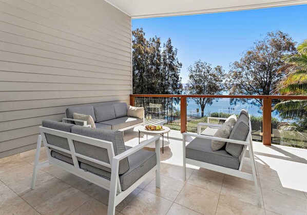 A modern outdoor seating area with a sofa and chairs, a small table with a fruit bowl, and a view of the sea and trees.