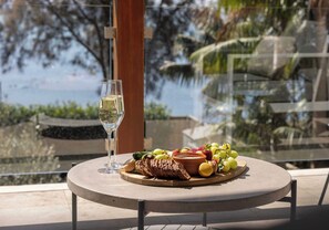 Ein runder Tisch mit einem Glas Weißwein, einer Speiseplatte mit Fleisch, Obst und einer Schüssel, sowie Blick auf ein Gewässer und Grünflächen.