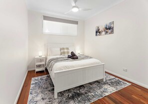 A bedroom with a bed, bedside tables, a ceiling fan, a window with blinds, a painting on the wall, and a patterned rug. - Esplanade Lake House, Pet Friendly (Oak Flats)
